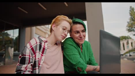 Close-up-shot-of-two-lesbian-girls-with-short-bright-haircuts-in-pink-and-green-checkered-shirts-looking-at-something-on-a-laptop-screen-while-sitting-on-a-small-step-near-a-building