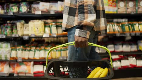 Side-view-of-a-man-with-Black-skin-color-and-wearing-a-plaid-shirt-and-blue-jeans-carries-a-grocery-basket-and-fills-it-with-selected-goods-during-his-shopping-in-a-grocery-supermarket