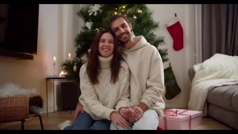 Portrait-of-a-happy-couple-guy-and-girl-in-white-sweaters-sitting-on-the-floor-near-a-pink-gift-hugging-and-looking-at-the-camera-in-a-cozy-Christmas-decorated-house-in-winter