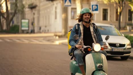 Happy-guy-with-long-curly-hair-in-a-green-helmet-and-a-denim-jacket-on-a-green-moped-with-a-flashlight-on-while-riding-in-a-summer-city