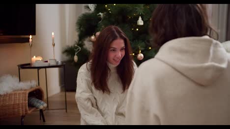 From-behind-the-shoulder,-a-brunette-guy-shakes-a-glass-ball-with-a-New-Year's-toy-and-then-shows-it-to-his-brunette-girlfriend-in-a-White-sweater-near-the-New-Year-tree-in-a-cozy-room-in-winter