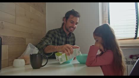 Happy-brunette-man-father-in-a-green-checkered-shirt-pours-milk-into-blue-plates-for-himself-and-his-daughter-brunette-girl-in-a-pink-dress-during-breakfast-at-the-table-in-the-kitchen