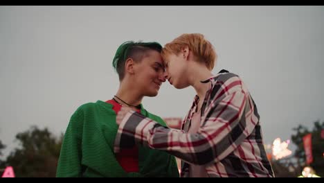 A-blonde-girl-with-short-hair-in-a-plaid-shirt-and-a-girl-with-green-hair-in-a-green-shirt-keep-their-faces-close-and-look-at-each-other-during-a-date-in-an-amusement-park