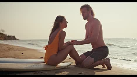 Side-view-of-a-blond-guy-with-a-bare-torso-in-green-shorts-holding-hands-with-his-blonde-girlfriend-in-an-orange-swimsuit-who-is-sitting-on-a-surfboard-on-a-sandy-rocky-beach-near-the-sea-in-the-morning