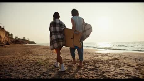 Rückansicht-Eines-Blonden-Mädchens-Mit-Grünem-Hut-Und-Kariertem-Hemd-Und-Ihres-Brünetten-Freundes-In-Weißem-T-Shirt,-Die-Am-Sandstrand-Entlang-Des-Meeres-Spazieren-Und-Morgens-Bei-Sonnenaufgang-Surfbretter-Tragen