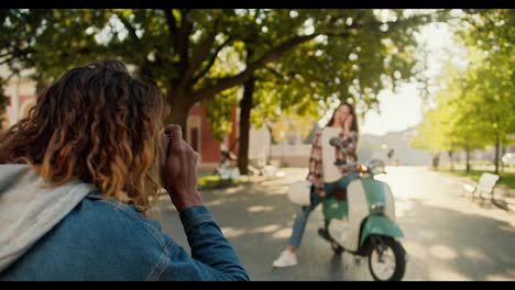 Over-the-shoulder,-a-guy-with-curly-hair-in-a-denim-shirt-takes-a-photo-of-his-brunette-girlfriend-in-a-checkered-shirt-and-a-white-T-shirt-near-a-green-moped-and-shows-her-a-like-sign-because-the-shot-was-a-success-in-a-beautiful-summer-city-park