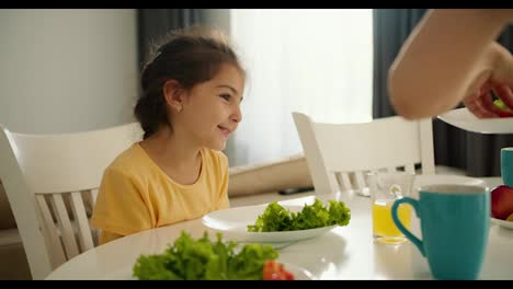 A-little-brunette-girl-in-a-yellow-dress-sits-on-a-chair-in-the-kitchen-and-her-mother-puts-green-herbs-and-vegetables-on-the-girl-s-plate-for-breakfast-at-the-table-in-the-morning