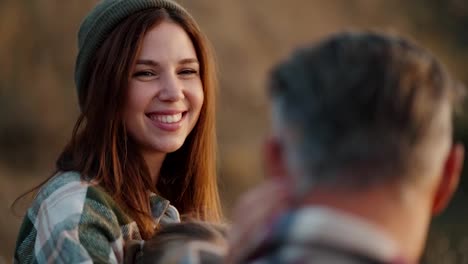 Over-the-shoulder:-A-happy-girl-in-a-green-hat-and-a-plaid-shirt-strokes-her-husband,-a-middle-aged-man-with-gray-hair,-during-her-vacation-outside-the-city-on-a-picnic-in-the-summer-evening