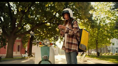 Una-Mensajera-Morena-Con-Una-Camisa-A-Cuadros,-Un-Casco-De-Motocicleta-Blanco-Y-Una-Bolsa-Amarilla-Está-Parada-Cerca-De-Su-Ciclomotor,-Mira-La-Pantalla-Del-Teléfono-Y-Alrededor-Para-Entender-Dónde-Entregar-Su-Pedido.