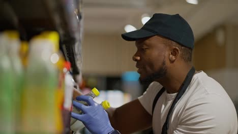 A-happy-man-with-Black-skin-in-a-black-cap-and-a-white-T-shirt-a-supermarket-employee-lays-out-and-sorts-bottles-in-a-special-display-case.-In-a-large-grocery-store