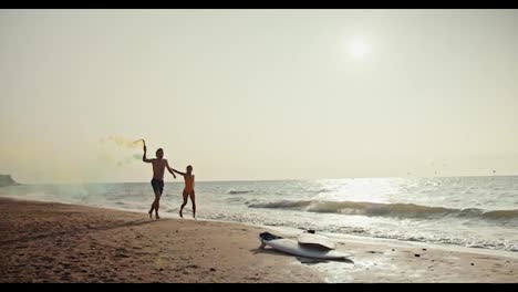 A-happy-couple,-a-blond-guy-in-shorts-and-his-blonde-girlfriend-in-an-orange-swimsuit-are-running-along-the-seashore-and-holding-yellow-and-green-fireworks-in-their-hands-that-leave-a-trail-of-smoke-in-the-morning-on-the-seashore