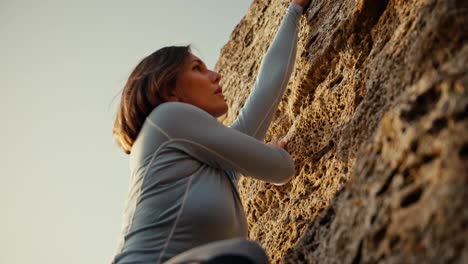 Shot-close-up,-shot-from-below,-a-blonde-girl-in-a-blue-jacket-climbs-a-steep-rock-on-which-the-rays-of-the-sun-fall.-blonde-girl-climber-climbs-up-a-steep-rocky-rock-and-looks-for-where-to-put-her-fingers-for-a-comfortable-spiral