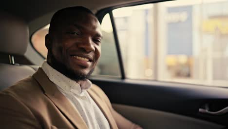 Portrait-of-a-happy-man-with-Black-skin-with-a-beard-in-a-brown-jacket-who-sits-in-the-back-seat-of-a-modern-car-smiling-and-looking-at-the-camera-during-his-business-trip-in-the-city