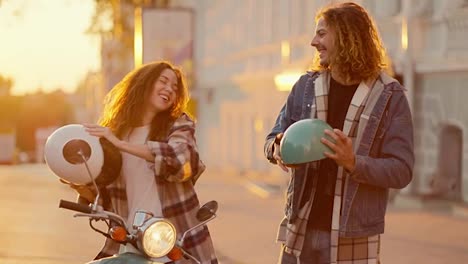 Happy-couple-on-a-golden-sunrise,-a-brunette-girl-sits-on-a-moped-in-a-plaid-shirt-near-her,-her-boyfriend-with-long-curly-hair-in-a-denim-jacket-hugs-her.-They-look-at-each-other-and-rejoice