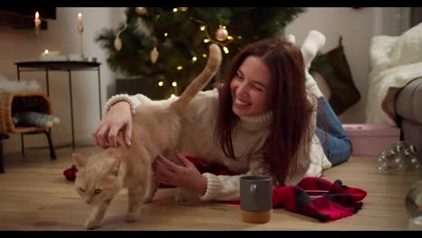 Una-Chica-Morena-Feliz-Con-Un-Suéter-Blanco-Se-Recuesta-En-El-Suelo-Sobre-Una-Manta-Roja-Y-Acaricia-A-Su-Gato-Color-Crema-En-Una-Acogedora-Habitación-Decorada-En-Estilo-Navideño.