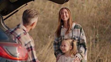 Close-up-shot-of-a-happy-brunette-girl-in-a-green-plaid-shirt-hugging-her-little-daughter-and-communicating-with-her-husband,-a-middle-aged-man-who-is-sitting-in-the-open-trunk-of-a-black-car-during-their-picnic-outside-the-city-in-the-summer