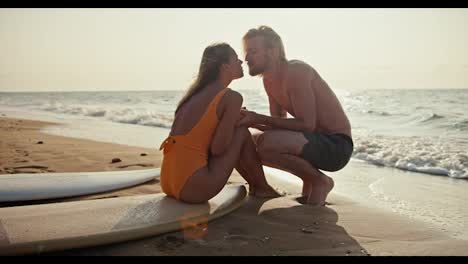 A-blonde-girl-in-an-orange-swimsuit-sits-on-a-surfboard,-communicates-with-her-blonde-boyfriend-and-kisses-him-on-the-sandy-beach-near-the-sea-in-the-morning-at-Sunrise