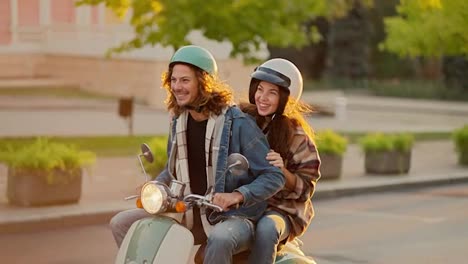 A-happy-couple,-a-guy-with-long-curly-hair-in-a-denim-jacket-and-a-green-moped-helmet,-rides-with-his-brunette-girlfriend-in-a-checkered-shirt-and-a-white-helmet-on-a-green-moped-along-a-green-summer-city-street