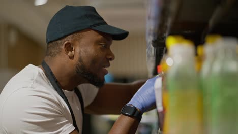 Filmado-De-Lado,-Un-Hombre-De-Piel-Negra-Y-Barba-Con-Una-Gorra-Negra-Y-Una-Camiseta-Blanca-Coloca-Y-Clasifica-Cosas-Y-Botellas-En-El-Mostrador-De-Un-Gran-Supermercado.