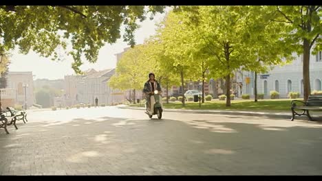 A-courier-guy-with-curly-hair-in-a-white-moped-helmet-and-a-denim-shirt-rides-his-green-moped-along-a-street-with-beautiful-green-trees-and-a-lawn-in-a-summer-city