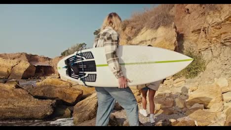 Rear-view-of-a-blond-guy-in-a-plaid-shirt-holding-a-white-surfboard-in-his-hands-and-walking-with-his-girlfriend-in-a-white-sweatshirt-who-is-also-holding-a-yellow-wooden-surfboard-on-a-sandy-rocky-beach-in-the-morning