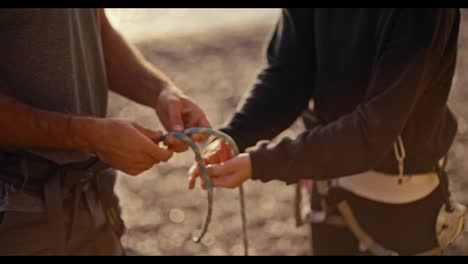 In-a-close-up-shot,-a-girl-in-a-black-gym-uniform-demonstrates-how-to-properly-tie-a-knot-with-a-thick-rope-on-a-guy's-equipment-and-belay-him-before-he-starts-climbing-a-rocky-cliff-during-a-rock-climbing-session.