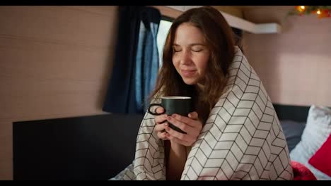 Close-up-shot-of-a-happy-brunette-girl-holding-a-black-mug-of-tea-in-her-hands-and-enjoying-the-aroma-and-warmth-of-the-drink,-wrapped-in-a-large-white-blanket-Sitting-in-a-trailer-in-a-camp-during-her-picnic-outside-the-city-in-the-summer