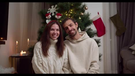 Portrait-of-a-happy-couple:-a-brunette-guy-in-a-white-sweater-and-a-brunette-girl-in-a-white-sweater-posing-and-looking-at-the-camera-near-their-New-Year-tree-in-a-cozy-winter-home