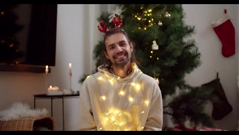 Portrait-of-a-happy-brunette-guy-in-a-white-sweater-who-looks-at-the-camera-and-smiles-in-a-New-Year's-yellow-glowing-garland-and-a-red-hoop-after-a-green-Christmas-tree-in-a-cozy-room-in-the-evening-in-winter