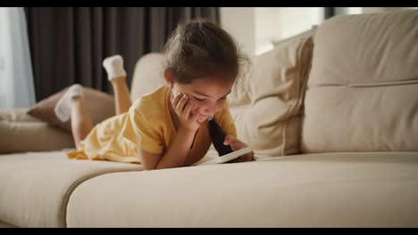 a-little-brunette-girl-lies-on-her-stomach-and-looks-at-the-screen-of-a-white-phone-on-a-light-brown-sofa-in-a-modern-apartment.-Little-girl-playing-a-game-or-watching-a-video-on-her-phone