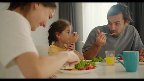 Un-Hombre-Moreno-Con-Barba-Incipiente-Desayuna-Con-Su-Esposa,-Que-Lleva-Una-Camiseta-Blanca,-Y-Su-Pequeña-Hija,-Que-Lleva-Un-Vestido-Amarillo,-En-La-Mesa-Familiar-Por-La-Mañana-En-Un-Apartamento-Moderno.