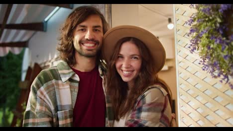 Close-up-portrait-of-a-happy-brunette-guy-with-stubble-and-a-plaid-shirt-and-a-red-T-shirt-who-stands-and-smiles-and-strokes-his-brunette-girlfriend-in-a-light-brown-hat-near-a-trailer-during-his-vacation-at-a-camp-during-a-picnic-outside-the-city-in-summer