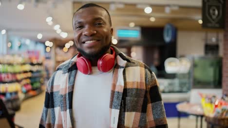 Portrait-of-a-confident-and-happy-man-with-Black-skin-in-a-plaid-shirt-and-red-wireless-headphones-walking-along-the-counters-in-a-modern-grocery-store