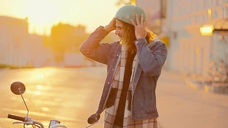 Portrait-of-a-Happy-guy-with-long-curly-hair-in-a-denim-shirt-who-takes-off-his-green-moped-helmet-from-his-head,-smiles-and-looks-at-the-camera-Near-his-moped-on-a-sunny-bright-morning-at-Sunrise