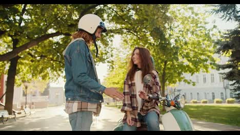 Glückliches-Paar:-Ein-Typ-Mit-Lockigem-Haar-In-Einer-Jeansjacke-Und-Einem-Weißen-Helm-Kommuniziert-Mit-Seiner-Brünetten-Freundin-In-Einem-Karierten-Hemd-Und-Einem-Weißen-T-Shirt,-Die-Auf-Einem-Grünen-Moped-In-Einem-Sommerlichen-Stadtpark-Sitzt