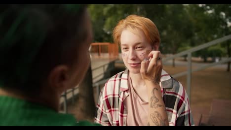 Over-the-shoulder:-A-girl-in-a-Green-shirt-with-short-straight-hair-wears-glitter-Yes,-the-cheeks-of-her-girlfriend-with-short-orange-hair-in-a-checkered-pink-shirt,-LGBT-couple-in-the-park-doing-makeup