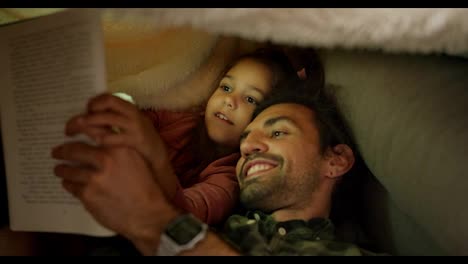 A-little-brunette-girl-in-a-pink-dress-lies-in-bed-with-her-father-who-tells-her-stories,-they-read-a-book-together-and-look-at-pictures-with-a-flashlight-on-near-a-gray-sofa-at-home