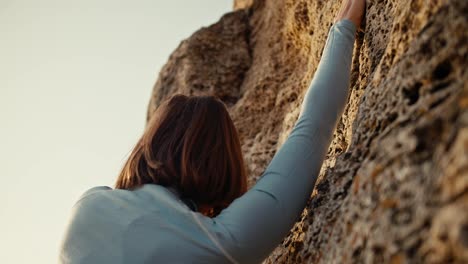 Close-up-shot-of-a-blonde-girl-with-a-bob-hairstyle-in-a-blue-jacket-climbing-a-steep-rock.-Girl-Climber-climbs-up-the-rock-and-looks-for-the-right-places-to-place-her-fingers-for-easy-climbing-on-the-rock