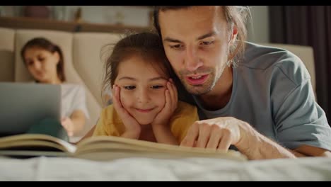 A-brunette-man-in-a-gray-t-shirt-is-reading-a-book-with-his-interested-little-brunette-daughter-in-a-yellow-t-shirt,-while-her-mother-is-relaxing-in-the-background-and-lying-on-the-sofa-with-a-laptop-in-a-modern-apartment