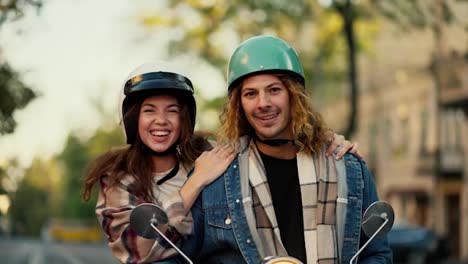 Portrait-of-a-happy-couple,-a-brunette-girl-in-a-plaid-shirt-hugs-her-boyfriend-with-long-curly-hair-in-a-denim-jacket-and-a-green-motorcycle-helmet-Near-her-motorcycle-in-the-summer-city