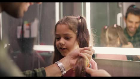 Happy-brunette-man-in-a-green-checkered-shirt-combs-his-little-daughter's-brunette-girl-hairstyle-with-a-ponytail-in-a-pink-dress-in-a-modern-bathroom