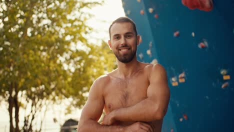 Retrato-De-Un-Escalador-Masculino-Feliz,-Un-Hombre-Moreno-Con-Barba-Incipiente-Cruza-Los-Brazos-Sobre-El-Pecho-Y-Posa-Cerca-De-Un-Muro-De-Escalada-Azul-En-Un-Día-Soleado-De-Verano.