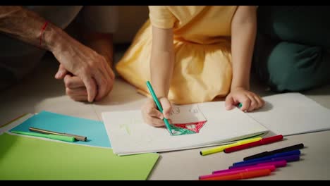 Close-up-shot-of-a-little-girl-in-a-yellow-dress-drawing-a-house-using-multi-colored-markers-on-a-piece-of-white-paper-while-sitting-on-the-floor-with-her-parents-in-a-modern-room