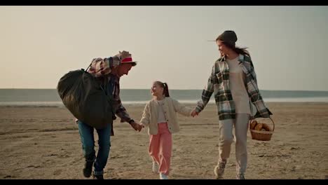 A-happy-brunette-man-with-gray-hair-in-a-checkered-shirt-carries-a-large-green-bag-and-walks-with-his-wife,-a-brunette-girl-in-a-Green-checkered-shirt,-and-with-his-little-daughter,-a-blonde-girl-in-a-White-jacket,-near-the-seashore-during-his-hike-and-picnic