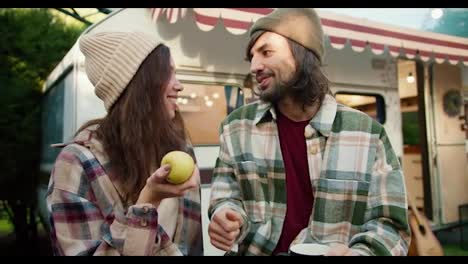 Close-up:-a-brunette-girl-holding-a-yellow-apple-in-her-hand-and-communicating-with-her-brunette-man-in-a-green-checkered-shirt-sitting-near-her-trailer-in-a-camp-during-a-picnic-outside-the-city-in-the-summer