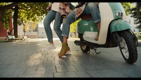 Close-up-shot-of-a-guy-in-jeans-sitting-on-a-green-moped-and-his-girlfriend-in-a-checkered-shirt,-jeans-and-white-sneakers-dancing-near-him-in-a-beautiful-summer-city-park