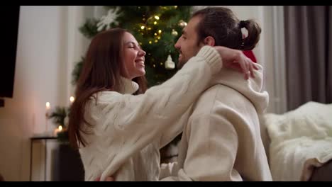 A-brunette-girl-in-a-white-sweater-and-a-guy-and-a-brunette-are-sitting-opposite-each-other-on-the-floor,-looking-at-each-other-and-the-guy-touching-the-girl-on-the-cheek-in-a-cozy-house-decorated-in-the-atmosphere-of-the-New-Year