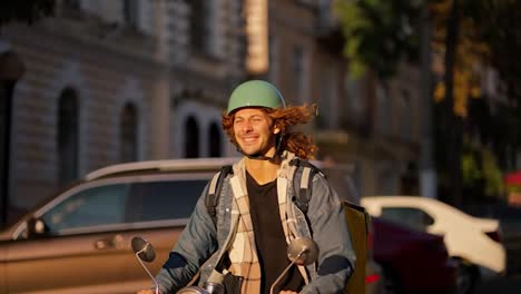 A-guy-with-long-curly-hair-in-a-denim-jacket-on-a-green-moped-rides-along-the-road-in-a-beautiful-summer-city