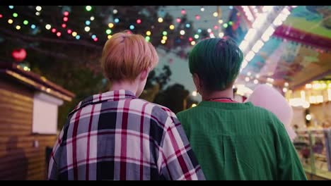 Rear-view-of-a-blonde-girl-with-a-short-haircut-in-a-plaid-shirt-and-a-girl-with-a-green-short-haircut-in-a-green-shirt-walk-through-a-bright-and-glowing-amusement-park-during-their-date