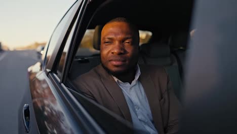 Close-up-shot-of-a-confident-man-with-Black-skin-color-and-a-beard-in-a-brown-suit-closes-the-rear-window-of-a-car-during-his-trip-in-a-modern-car